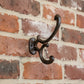 A rustic black cast iron coat rack hook mounted on a warm wood-paneled wall, with a softly blurred interior scene in the background showing a scarf on hooks and shoes on the floor.