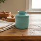 A light blue ceramic butter pail with a matching lid, featuring an embossed oval logo that reads 'Butter', set against a draped beige fabric background.